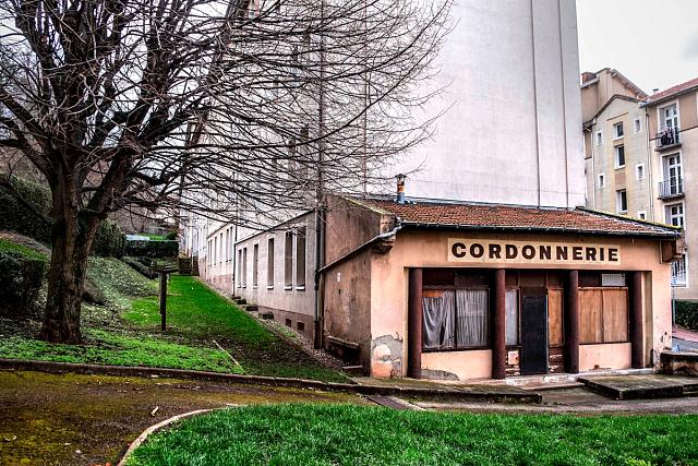 Une petite boutique de cordonnerie ancienne appuyée sur un immeuble moderne entourée d'une pelouse.