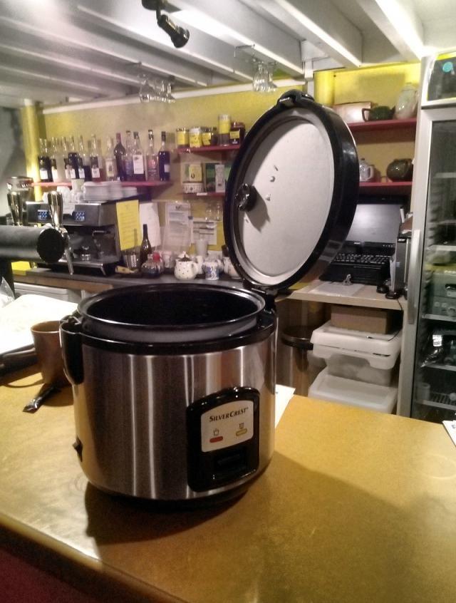 Sur un meuble de bar est posé une cuiseuse à riz ouverte. Derrière, du matériel de bar.